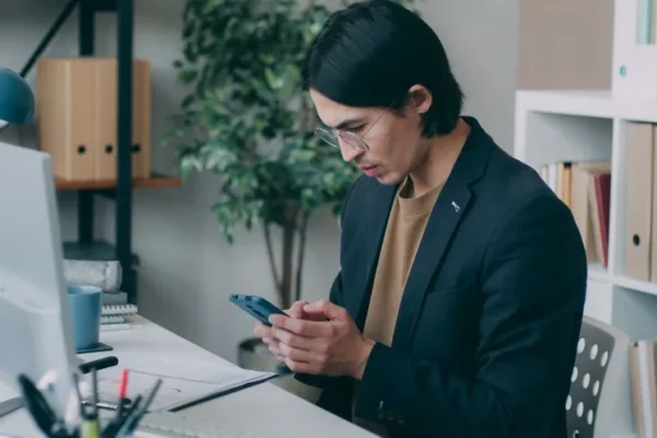 Young man in an office using smartphone while working at a computer desk.